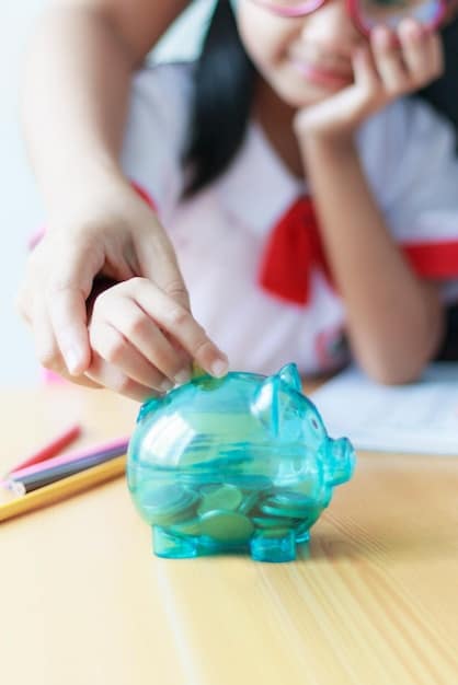 A close-up of a child's hand placing coins into a piggy bank, symbolizing saving money for future college expenses. The background is blurred, featuring textbooks and a calculator.